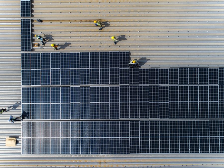 Luftaufnahme einer Gewerbehalle mit Trapezblechdach, auf der ein Montageteam Photovoltaikmodule verlegt